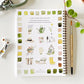 Spiral-bound notebook with watercolor illustrations of garden-themed designs on a white surface with art supplies.