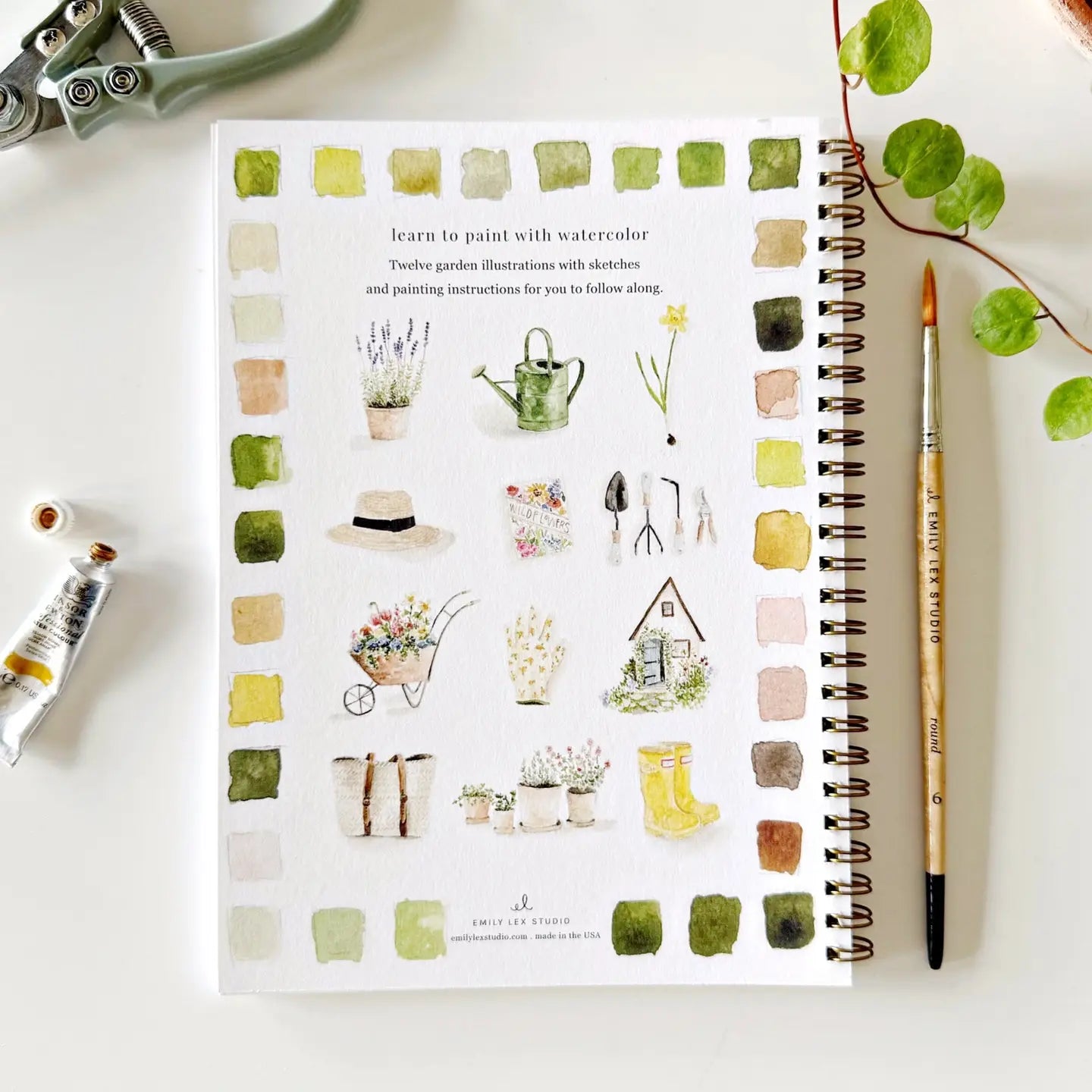 Spiral-bound notebook with watercolor illustrations of garden-themed designs on a white surface with art supplies.