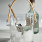 Clear glass ice bucket with a wooden handle and metal scoop, containing ice cubes.