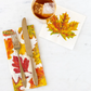 Table setting with fall-themed napkins, fork, knife, and cup of iced coffee on a white surface.