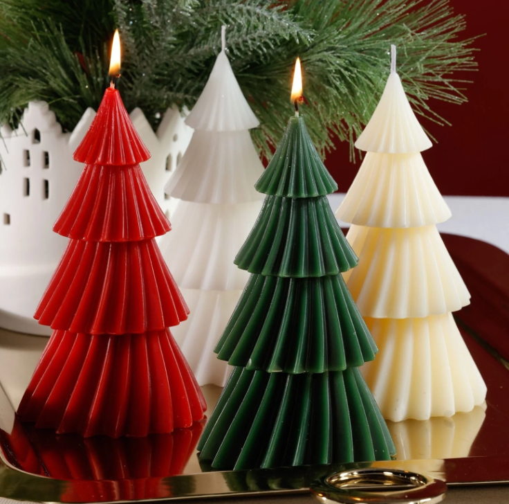 Three Christmas tree-shaped candles in red, green, and white on a reflective surface with a blurred background.
