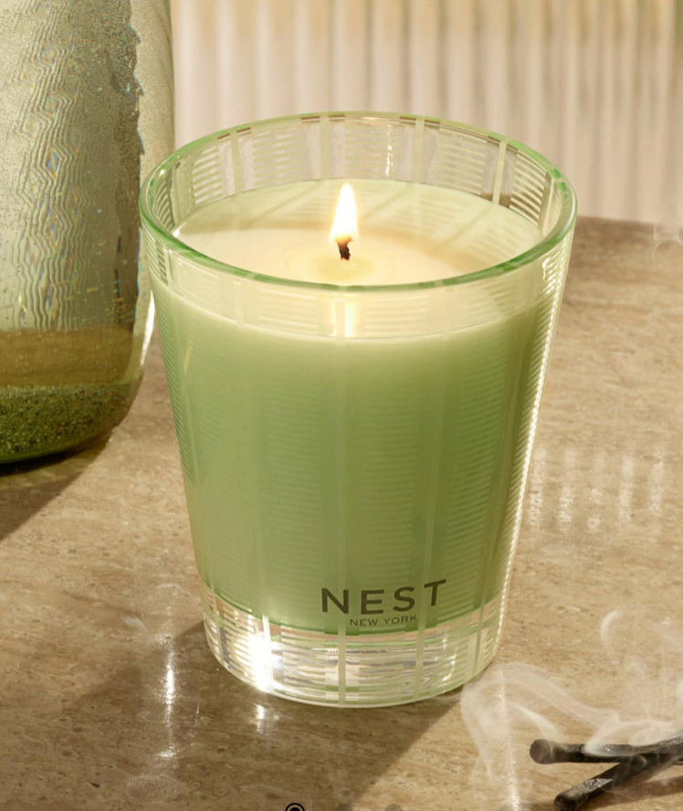 Green candle in a glass holder with 'NEST' branding on a textured surface.
