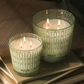 Two lit candles in green patterned containers on a surface with a striped background