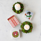 Two small green wreath candle holders with white candles, a box of matches, and a small card with a snowman design on a marble surface.