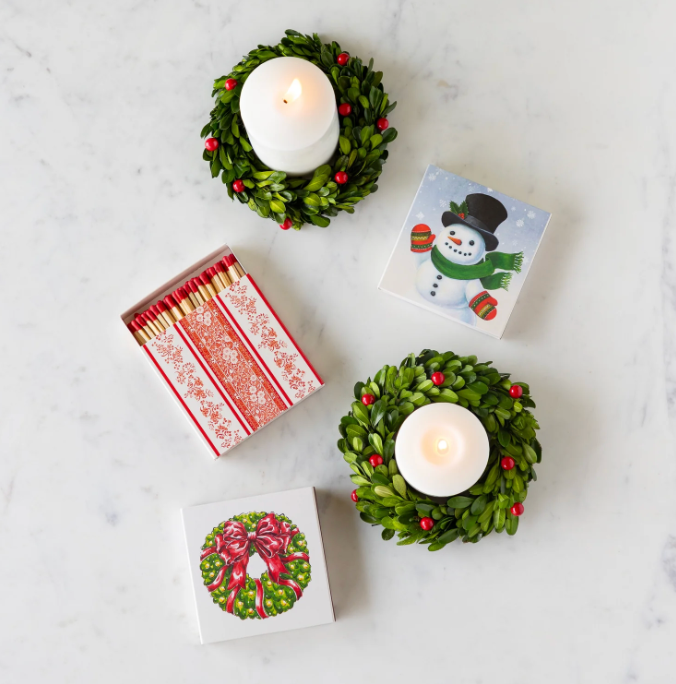 Two small green wreath candle holders with white candles, a box of matches, and a small card with a snowman design on a marble surface.